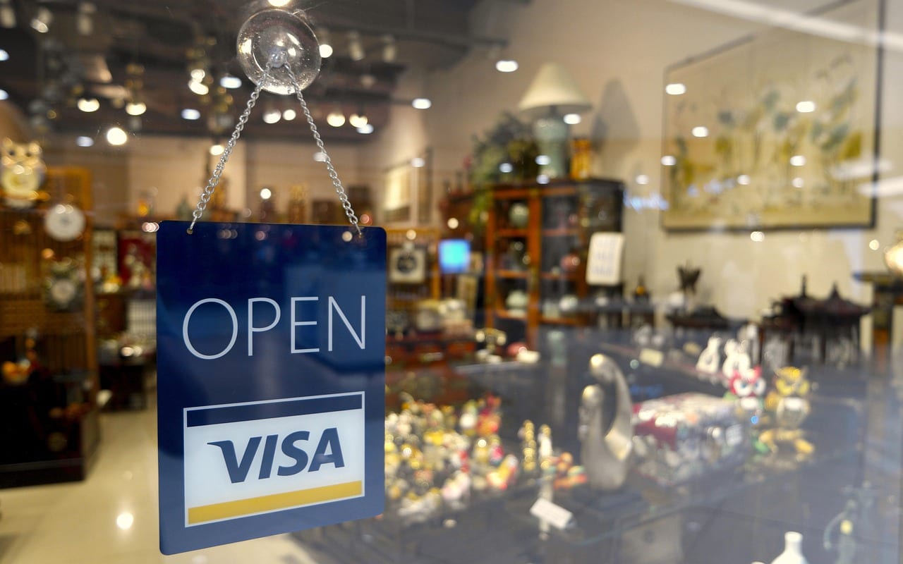 Reflection in a shop window displaying an 'OPEN' sign with a Visa logo; the interior is warmly lit, showcasing shelves and a variety of goods.