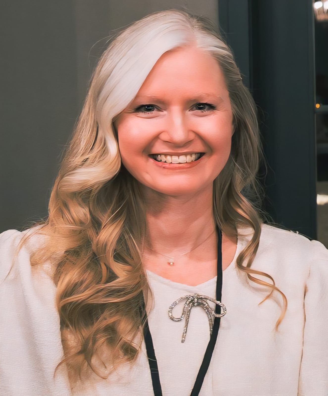 Portrait of a smiling woman with long, wavy light hair wearing a cream-colored blouse and a lanyard, suitable for business or organizational representation